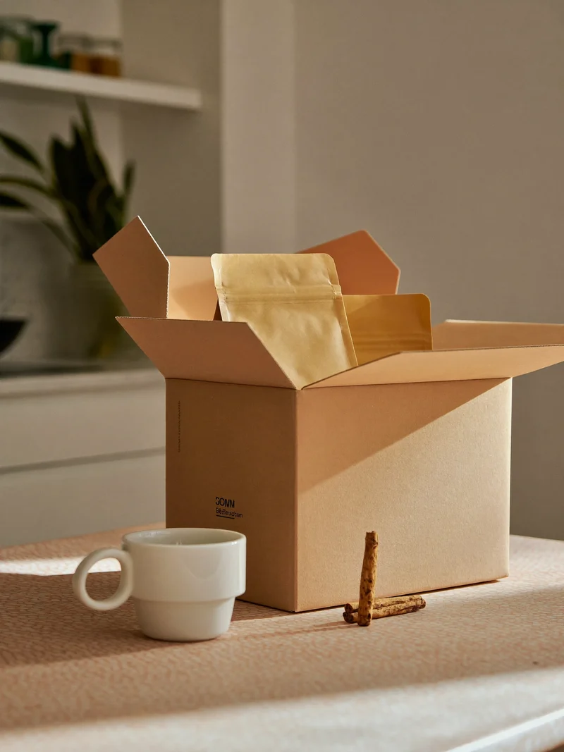 Geöffnete Lieferbox mit unbeschrifteten Kaffeetüten, Keramiktasse und Zimtstange auf einem Küchentisch